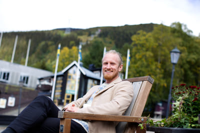 Fredrik Brännlund, founder of Noa The Game Fredrik Brännlund sitting on a wooden bench outdoors in a beige coat, a forested mountainside behind him